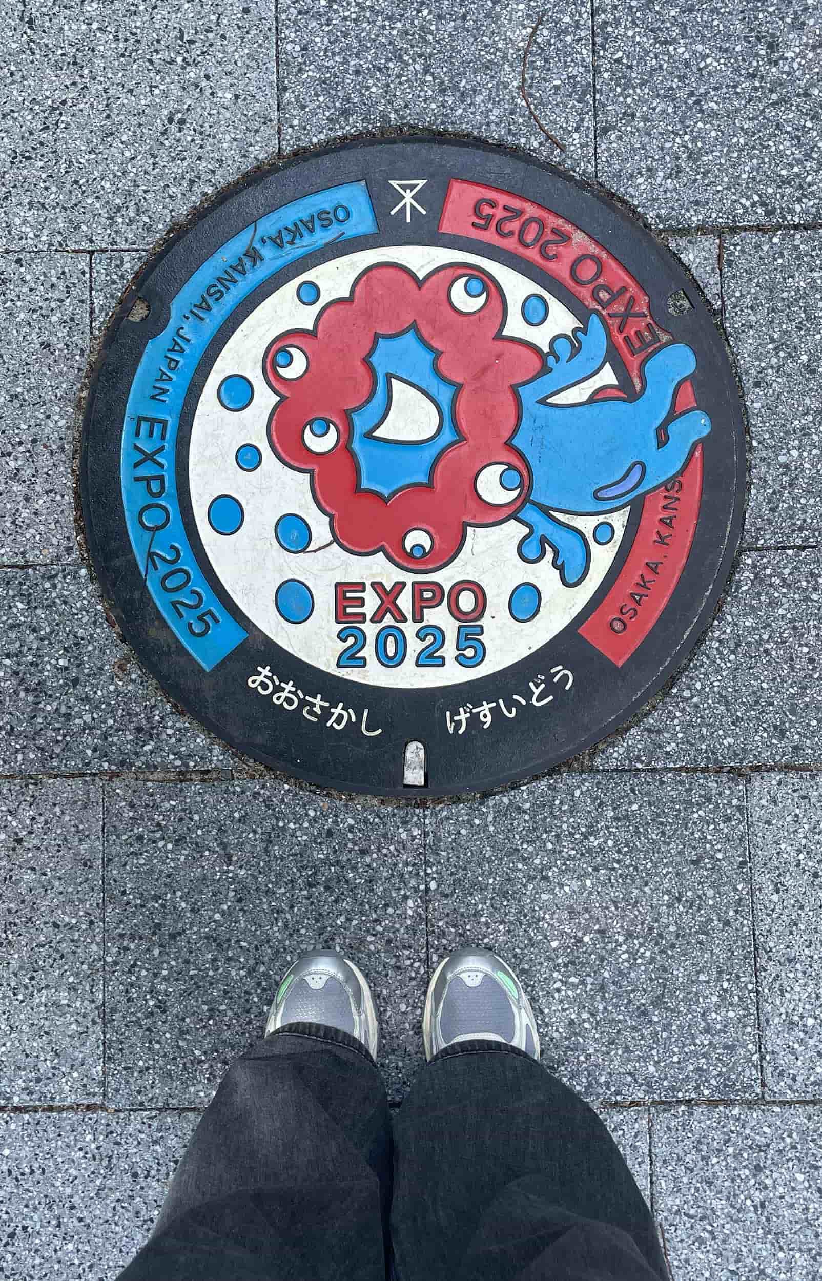 Snapshot from above showing a person’s feet near the Expo 2025 Osaka, Japan logo embedded in the street surface.