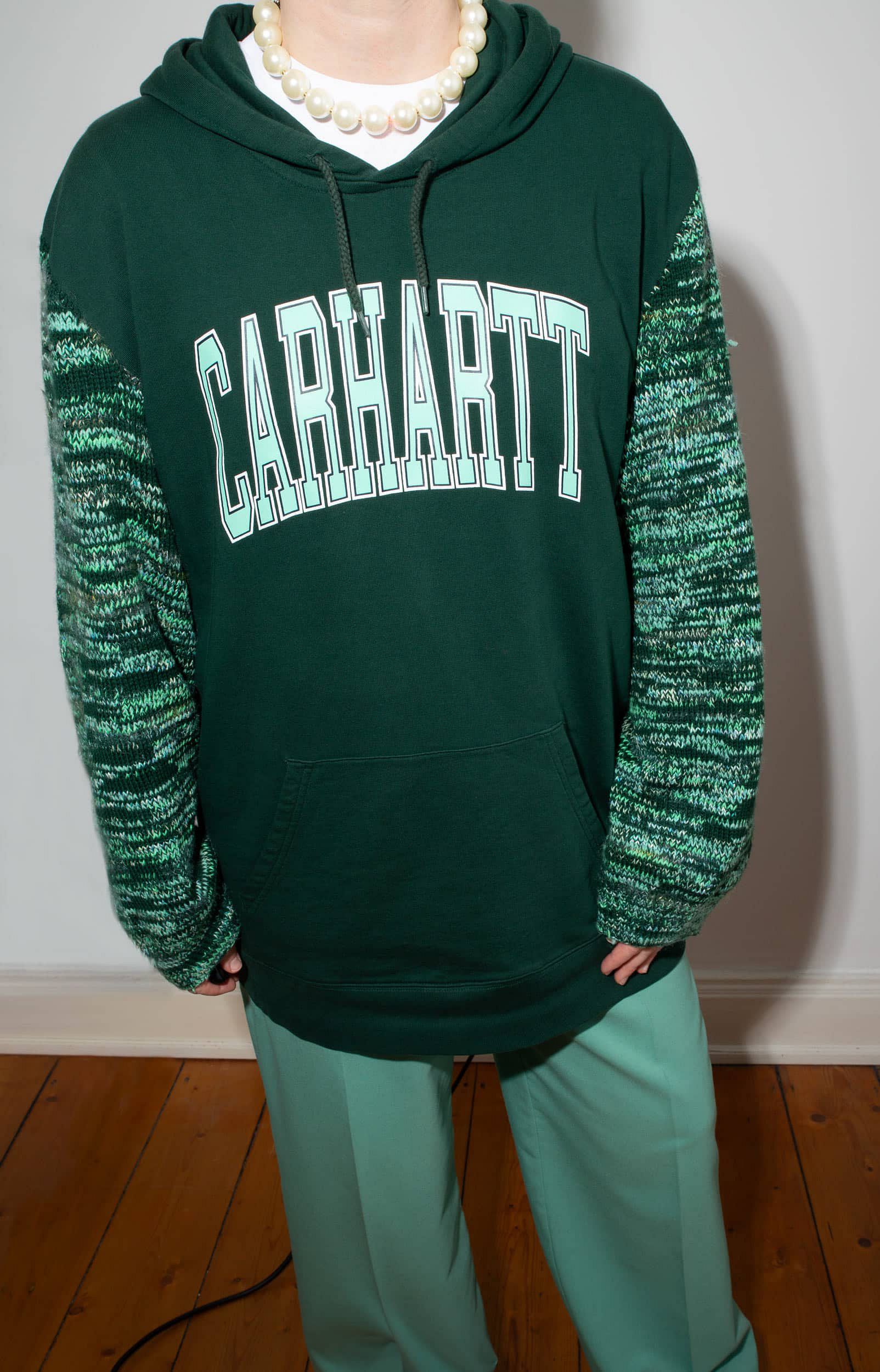 Woman wearing a dark green KnitInProgress sweater with sleeves knitted from various leftover yarns, paired with green pants in a different shade, accessorized with a chunky beaded necklace. The head is cropped out of the frame.