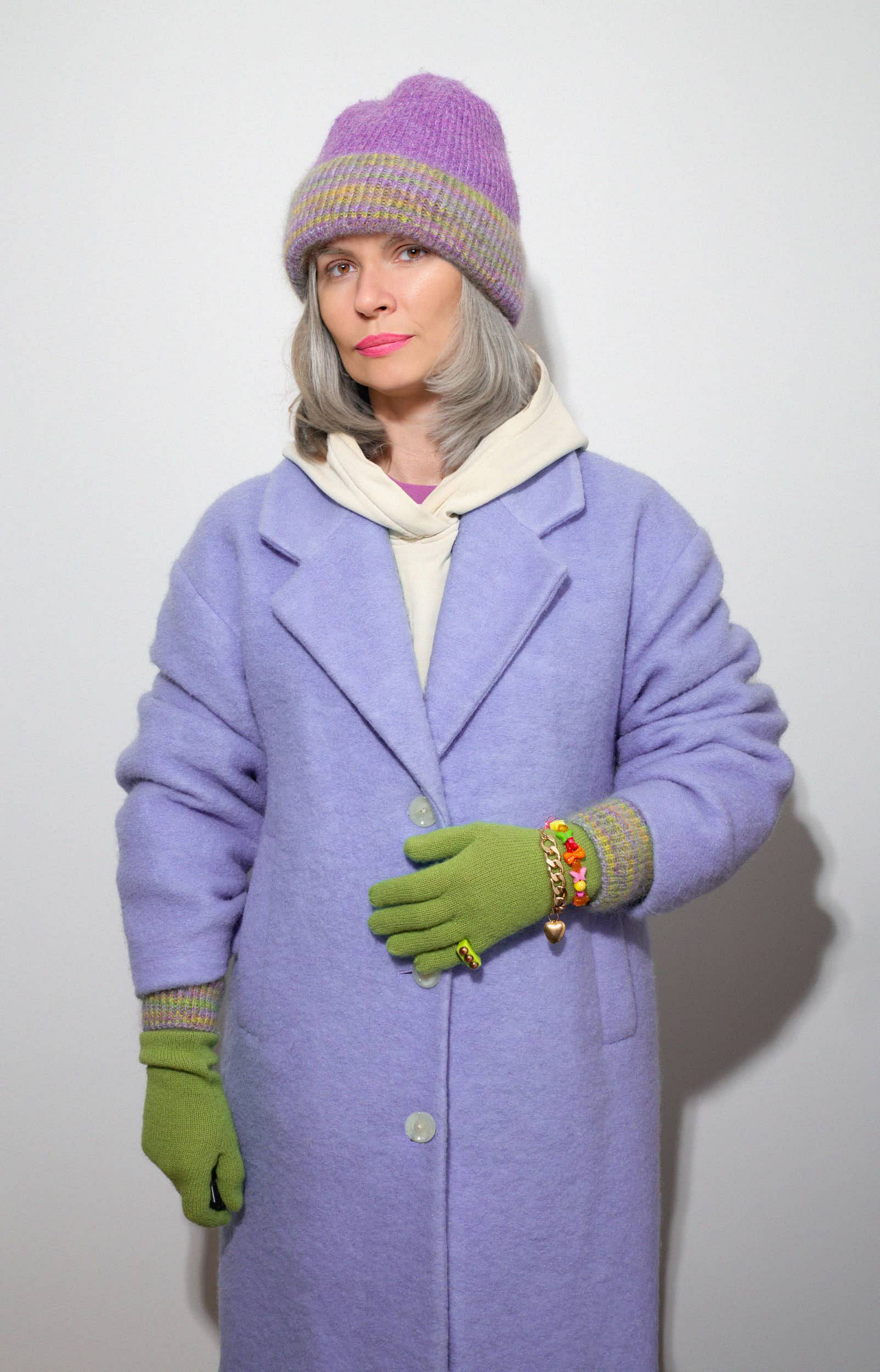 Woman wearing the green and purple set from the MultiColourCombo collection, including matching hat and gloves with color-coordinated cuffs, paired with a light blue winter coat, casual hoodie, and accessories including bracelets and rings.