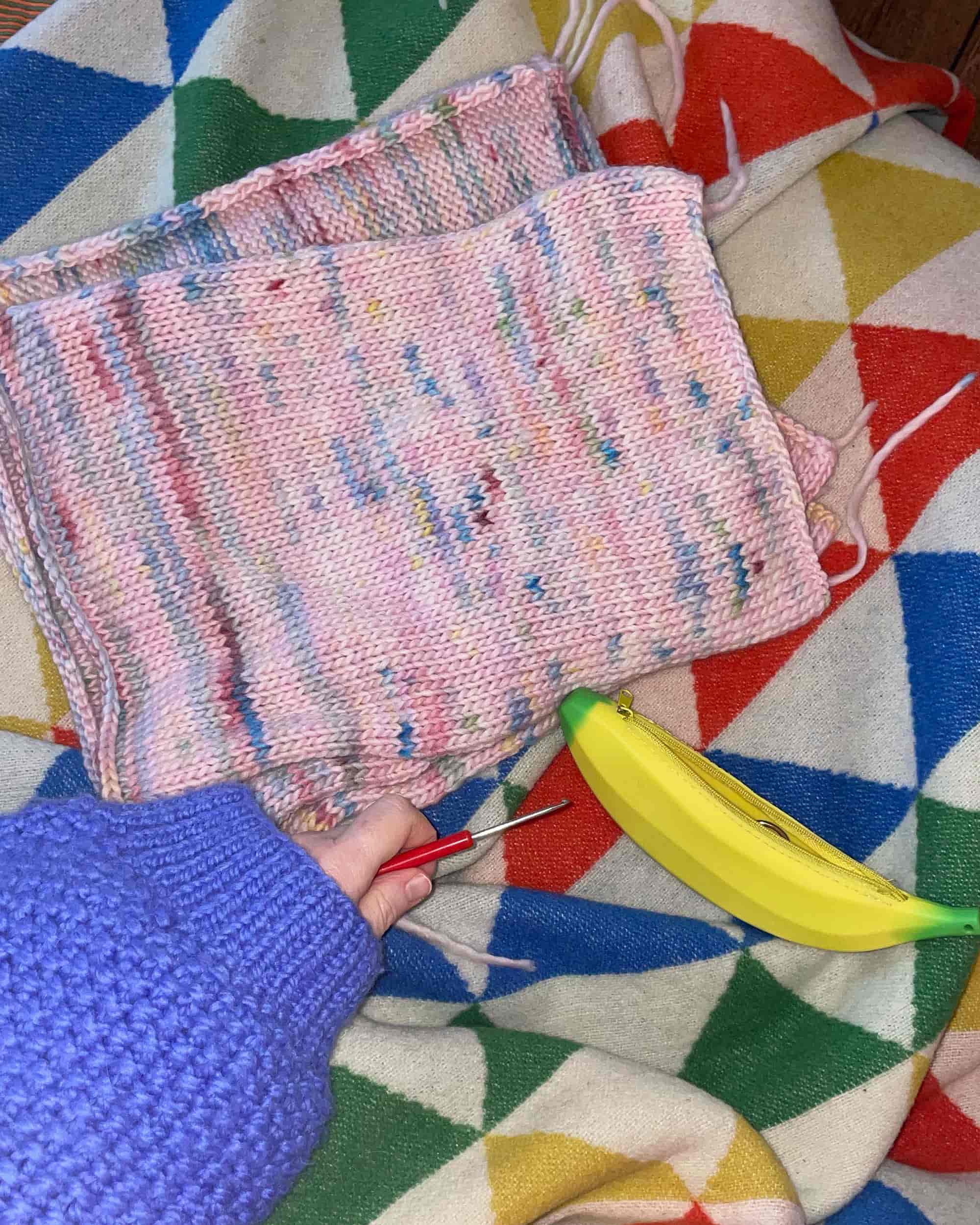 Top view of a person in a blue cardigan holding a crochet hook, surrounded by finished pieces that will later form the 'NextToYou' project.