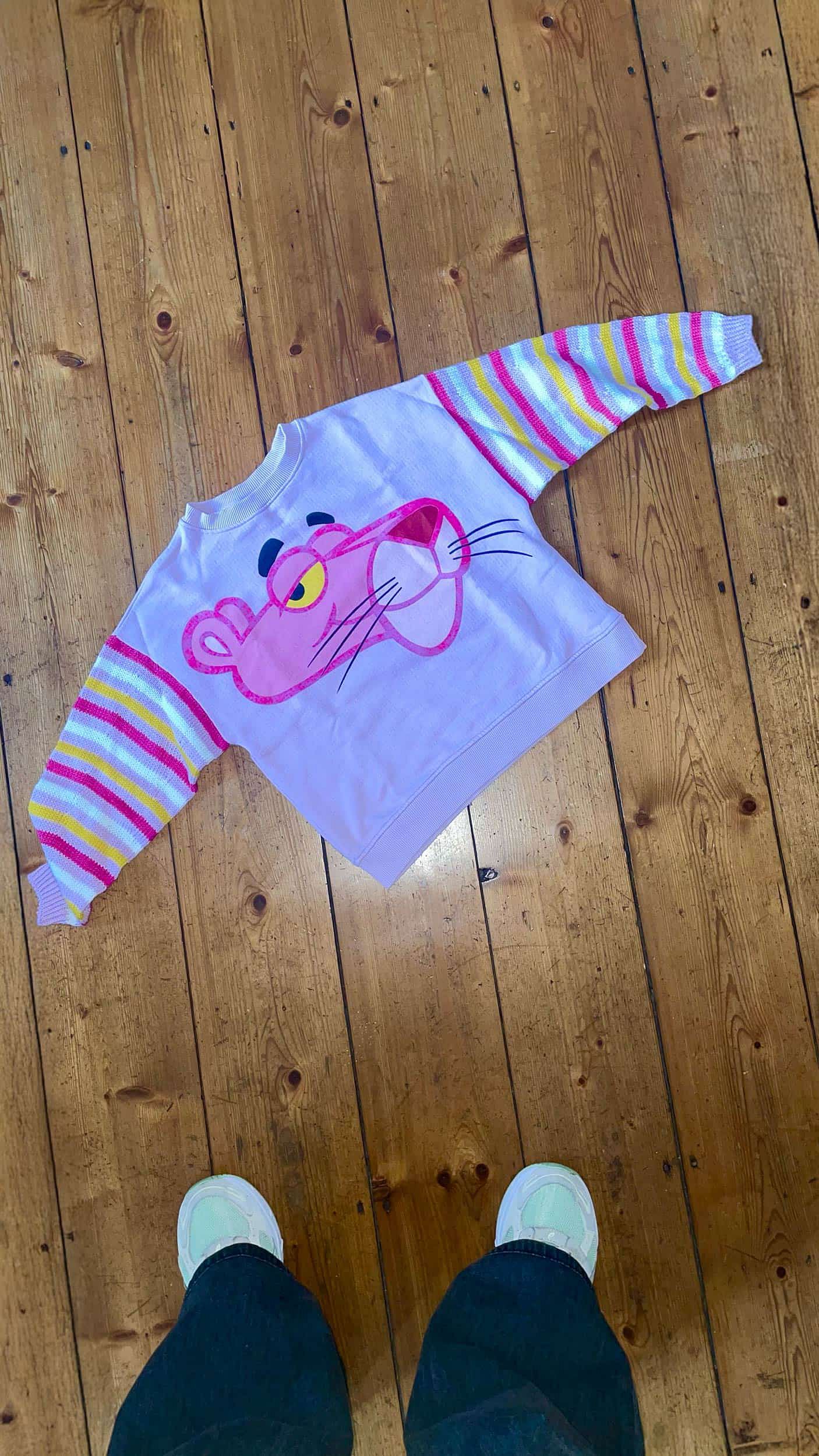 Top-down view of a finished KnitInProgress sweater with a Pink Panther motif, laid out on a wooden floor. Sleeves are knitted in pink, white, and yellow to match the design. Feet of the person taking the photo are partially visible.