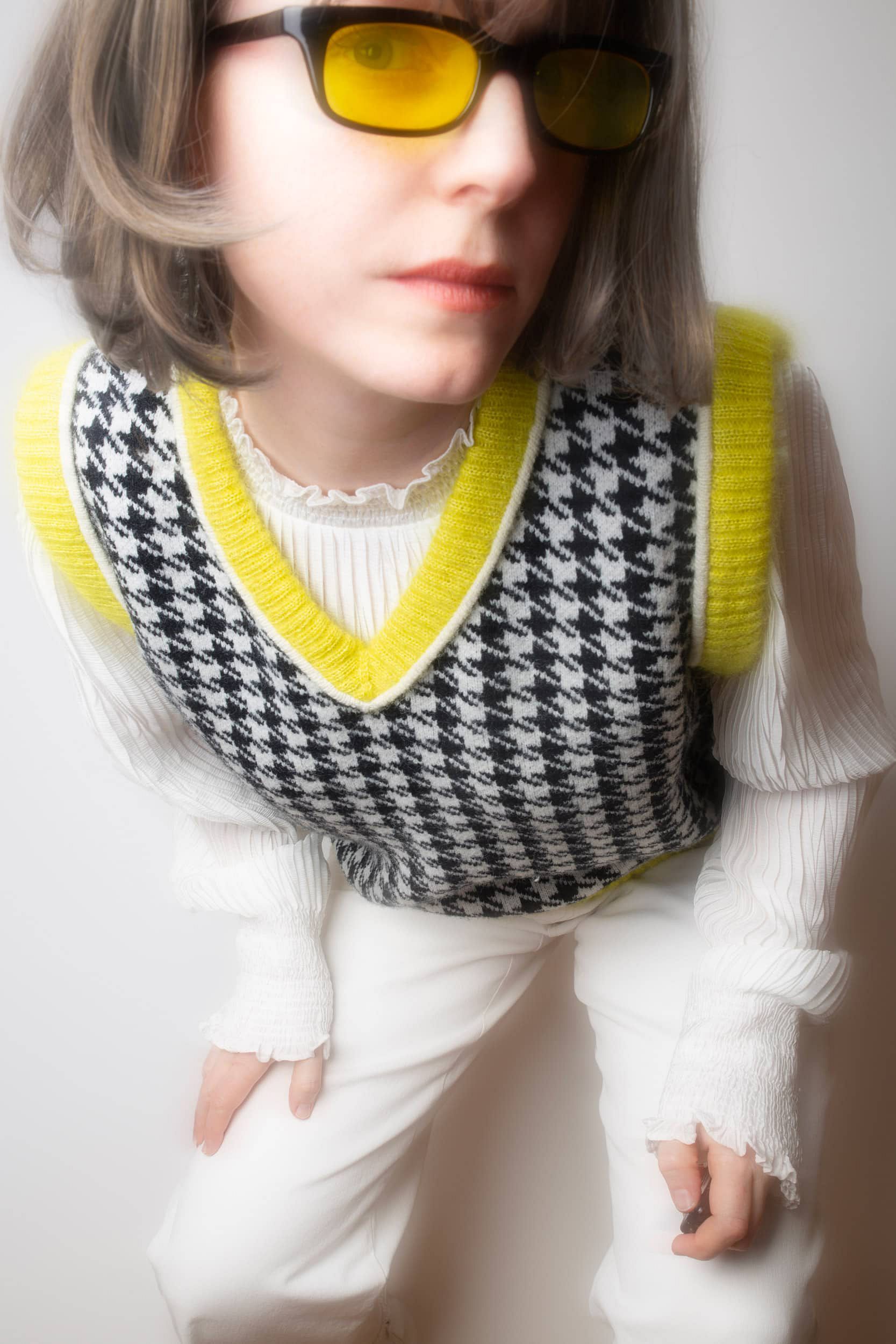 Person leaning toward the camera, wearing a reworked black-and-white houndstooth vest by Next Level Knit with colorful new ribbing. The full outfit is white, styled with yellow-tinted sunglasses matching the updated trim.