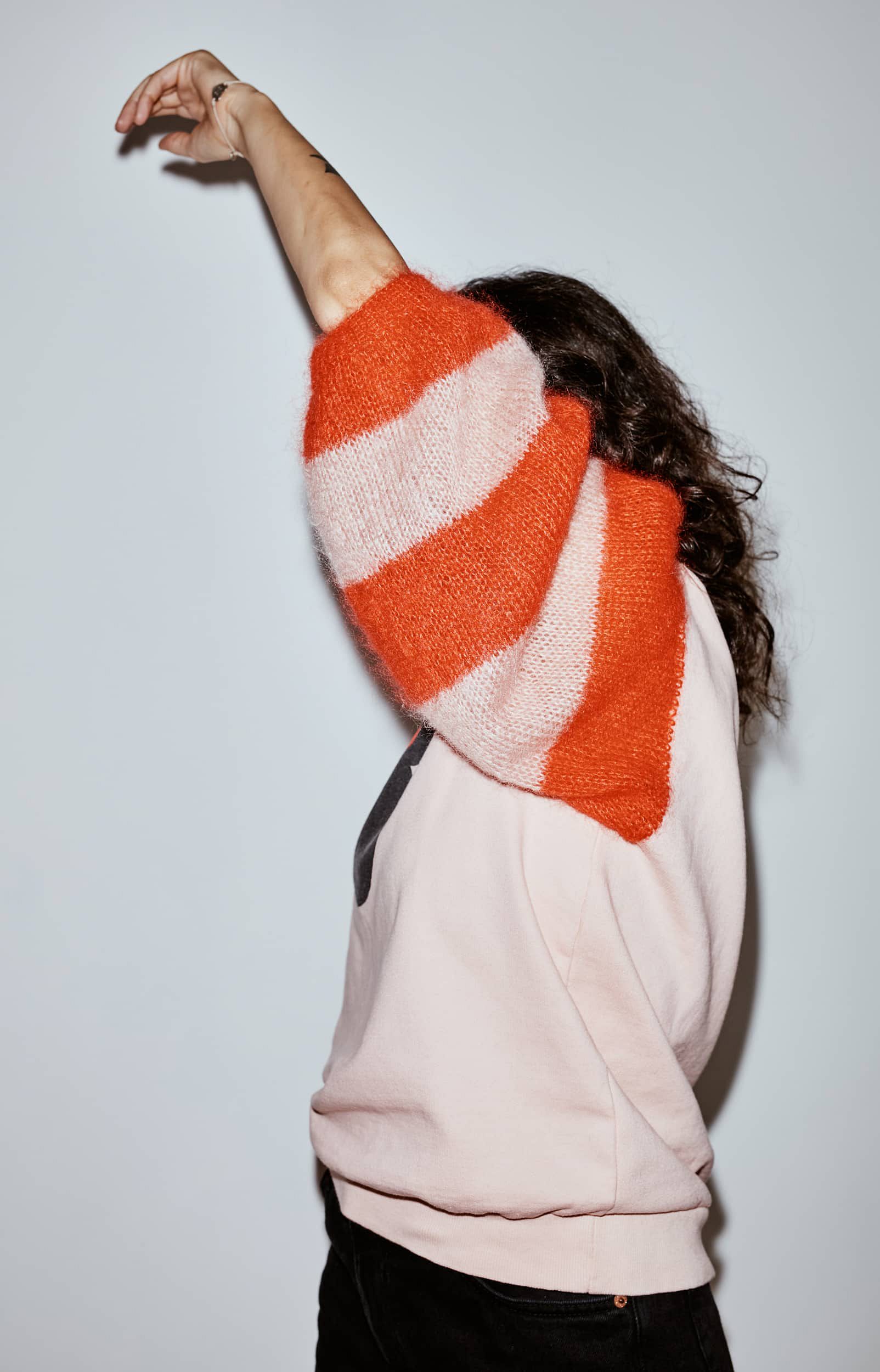 Portrait of a woman with curly hair wearing a pink sweatshirt with a Minnie Mouse print and eye-catching red and pink knitted sleeves - photographed against a light-coloured background.